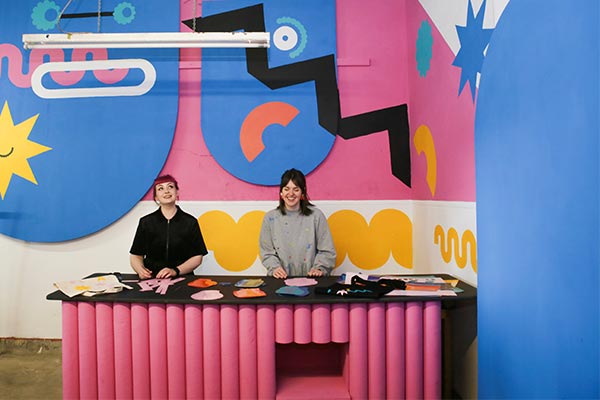 Two women stand behind a bench in a room filled with vibrant graphic shapes.