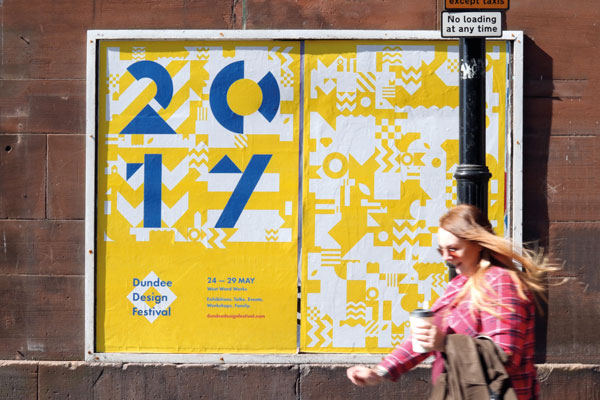 Woman with coffee in hand passes festival posters in the street