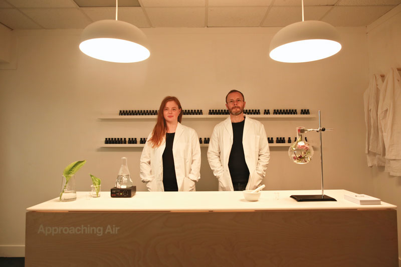 Man and woman stand side by side softly lit in the scent lab space. Various equipment and vials fill the scene.