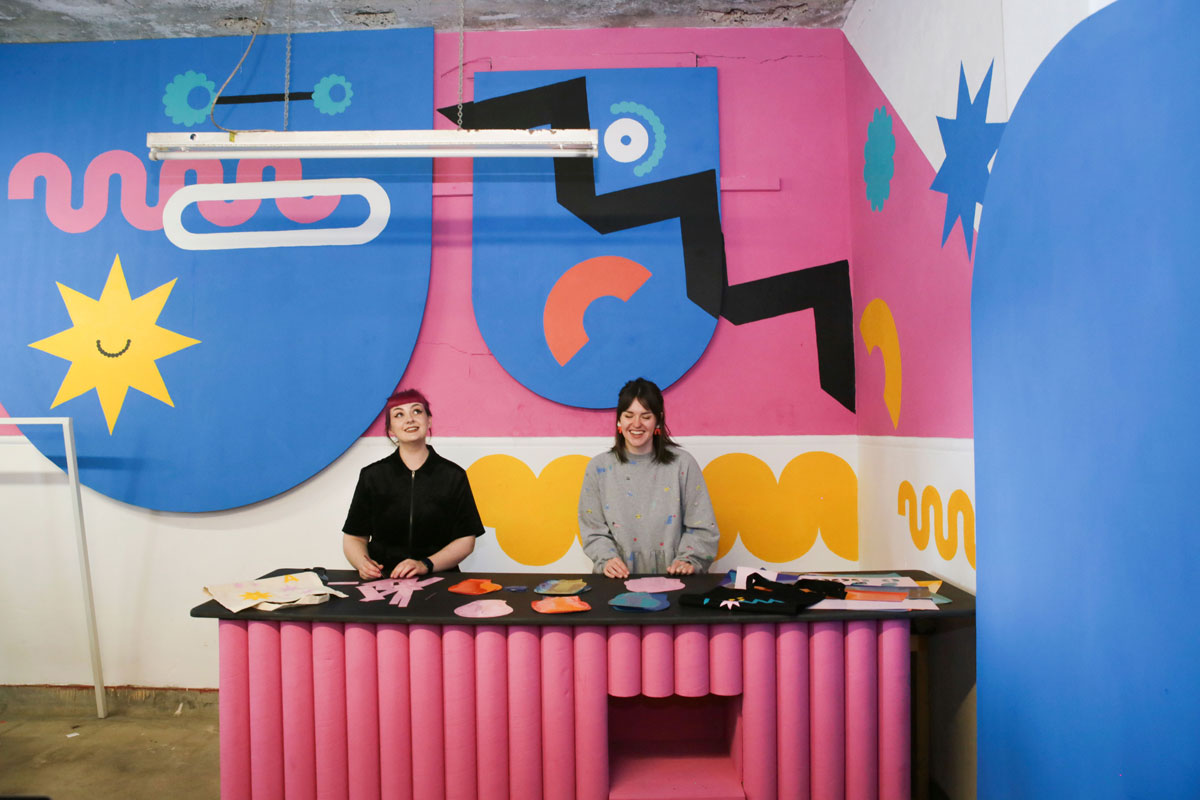 Two women stand behind a bench in a room filled with vibrant graphic shapes.