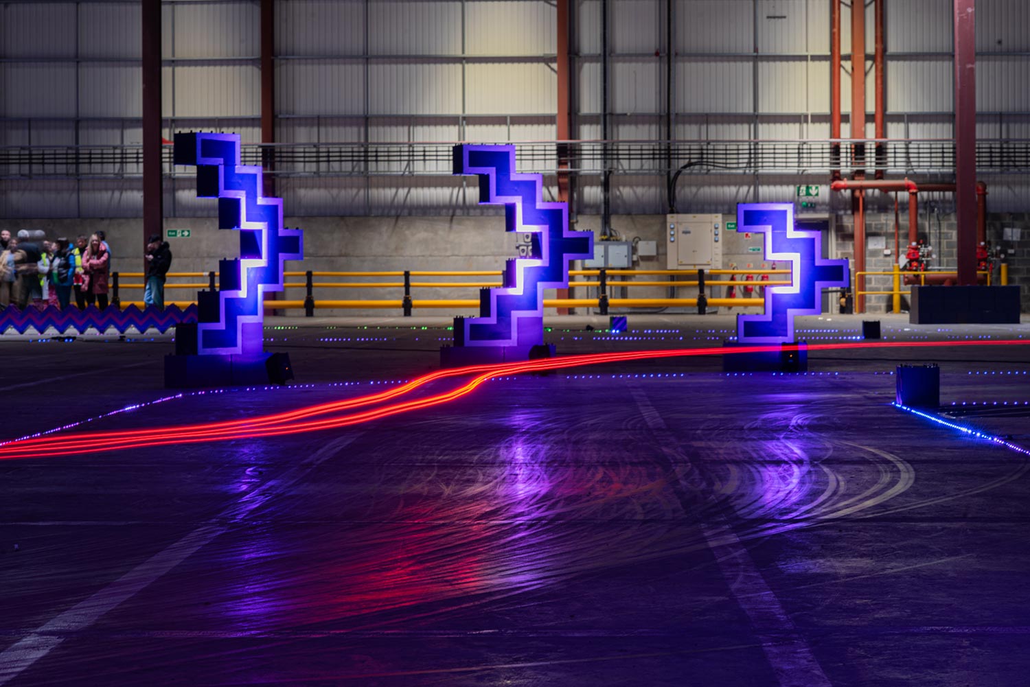 Light streaks from go-kart tail lights swoop past on the corner of a purple illumintaed racetrack.