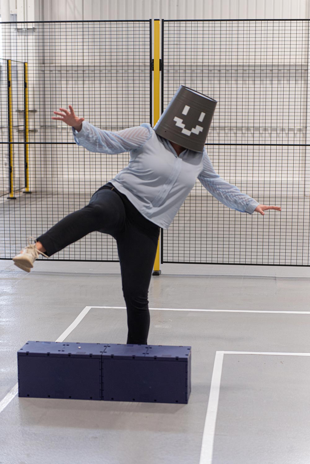 Woman with bucket on head makes silly pose stepping over block.