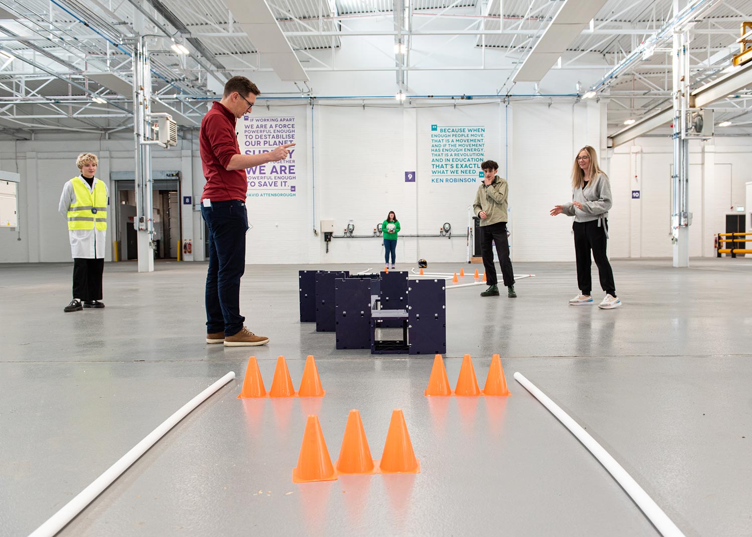3 people in a large white warehouse space point and gesture as they try to naviagte a track of blocks and cones.