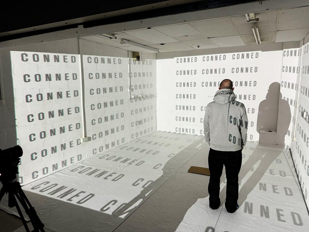 A man in a hoodie stands in a white room with the word conned projected in rows across the walls and floor.