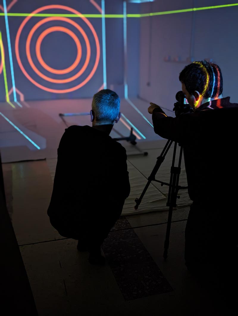 Two men beside a tripod point into a room being projection mapped.